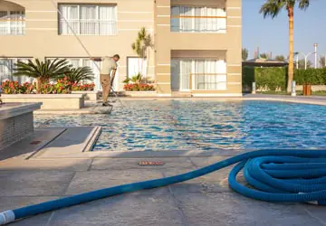 Pool maintenacne worker skimming pool with pool vaccuum tub in foreground